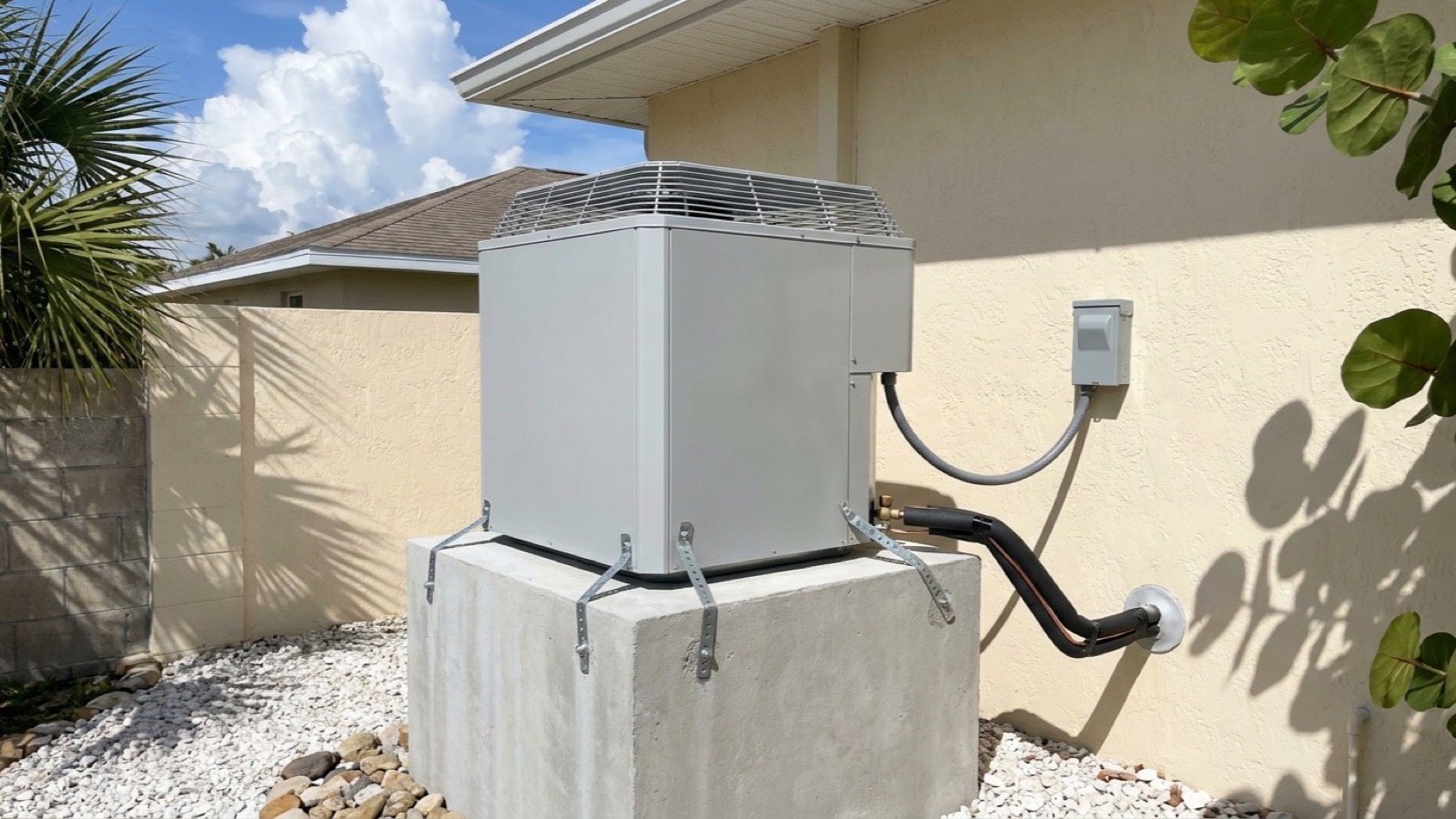 An outdoor air conditioning condenser raised on a tall poured concrete stand beside a low block and stucco Englewood home, galvanized tie-down straps running over the cabinet base, crushed shell and sabal palms around the pad