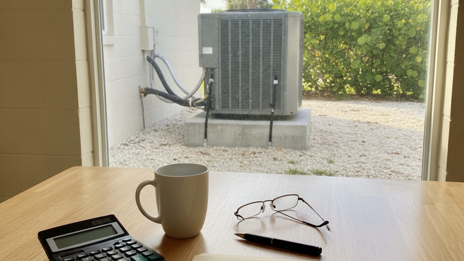 A kitchen table in a single story Englewood Florida block and stucco home, a calculator, coffee mug, reading glasses and a pen beside a closed folder, a new outdoor AC condenser on an elevated pad with hurricane tie down straps visible through the window, warm Gulf coast morning light