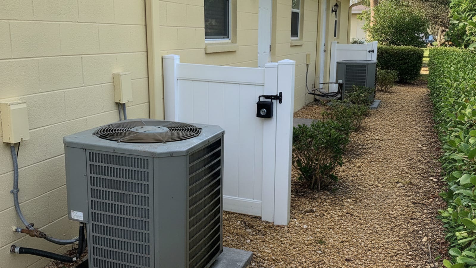 An older outdoor air conditioning condenser on a cracked concrete pad in the side yard of a pale yellow block and stucco Englewood rental duplex, a lockbox hanging on the gate latch, crushed shell driveway and sabal palms, bright Gulf coast morning light